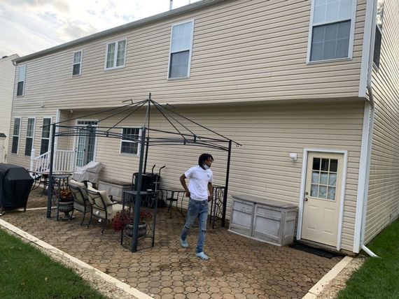 A person walks on a patio in front of a two-story beige house. The patio has a black canopy and outdoor furniture.