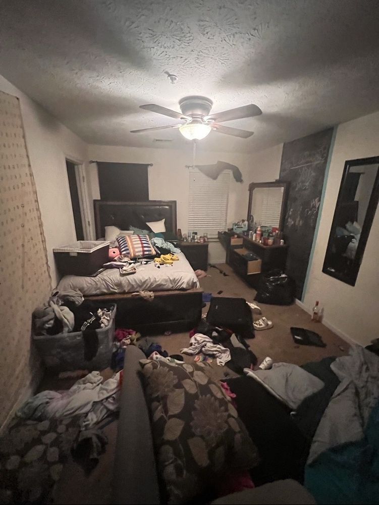 Messy bedroom with unmade bed, clothes scattered, and furniture. Poor lighting.