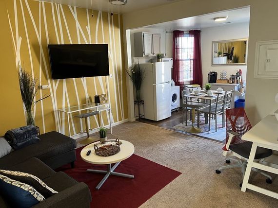 Cozy living space with a dark gray sectional, TV on a yellow accent wall, and open kitchen with dining area.