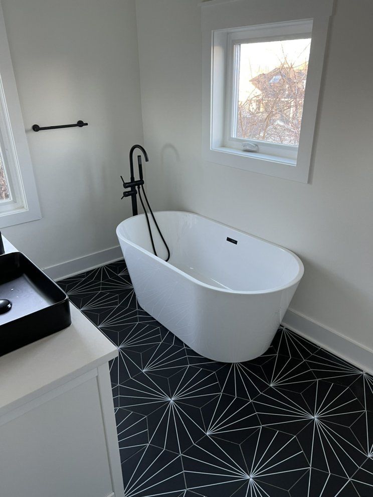 Modern bathroom with a white oval bathtub, black geometric floor tiles, and black fixtures. A window provides natural light.