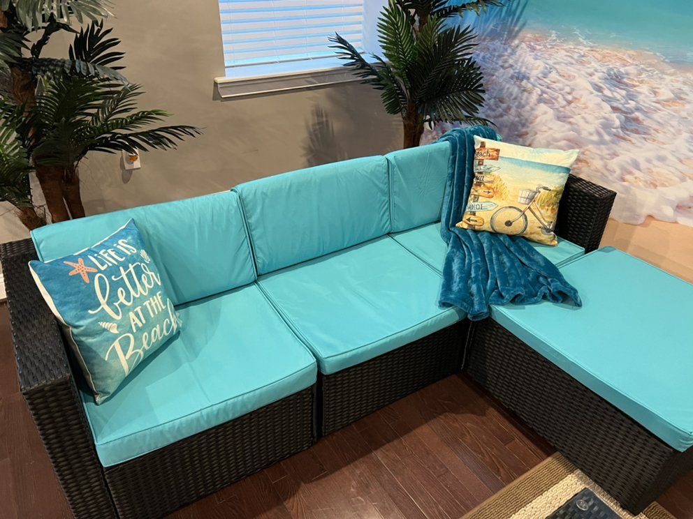 Wicker sectional sofa with turquoise cushions and throw pillows, set in a beach-themed room with artificial palm trees.