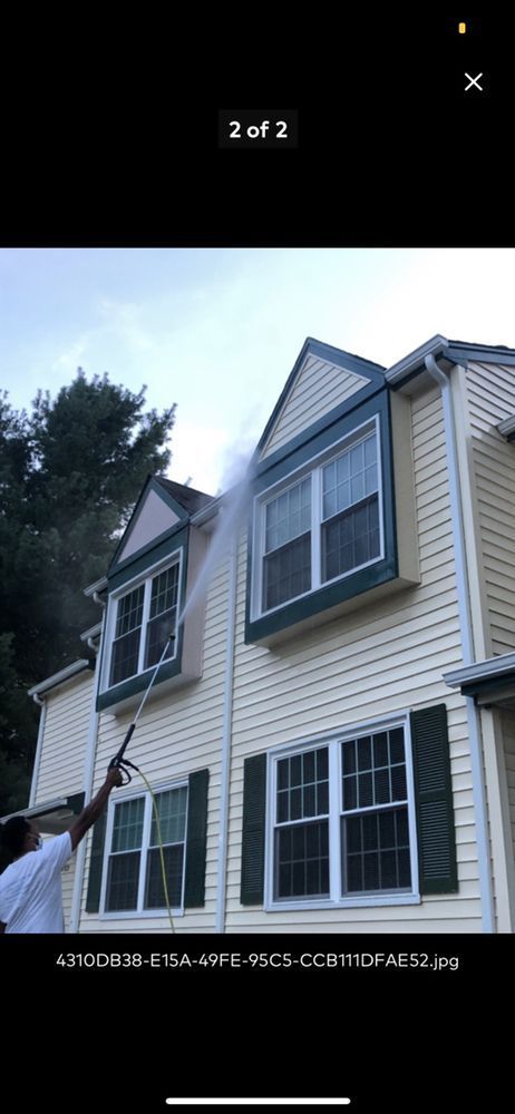 Person power washing the exterior of a two-story beige house with dark green shutters and trim.