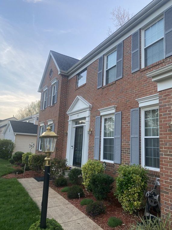 Two-story brick house with grey shutters, a black front door, and a lamp post. Green bushes and red mulch border the walkway.