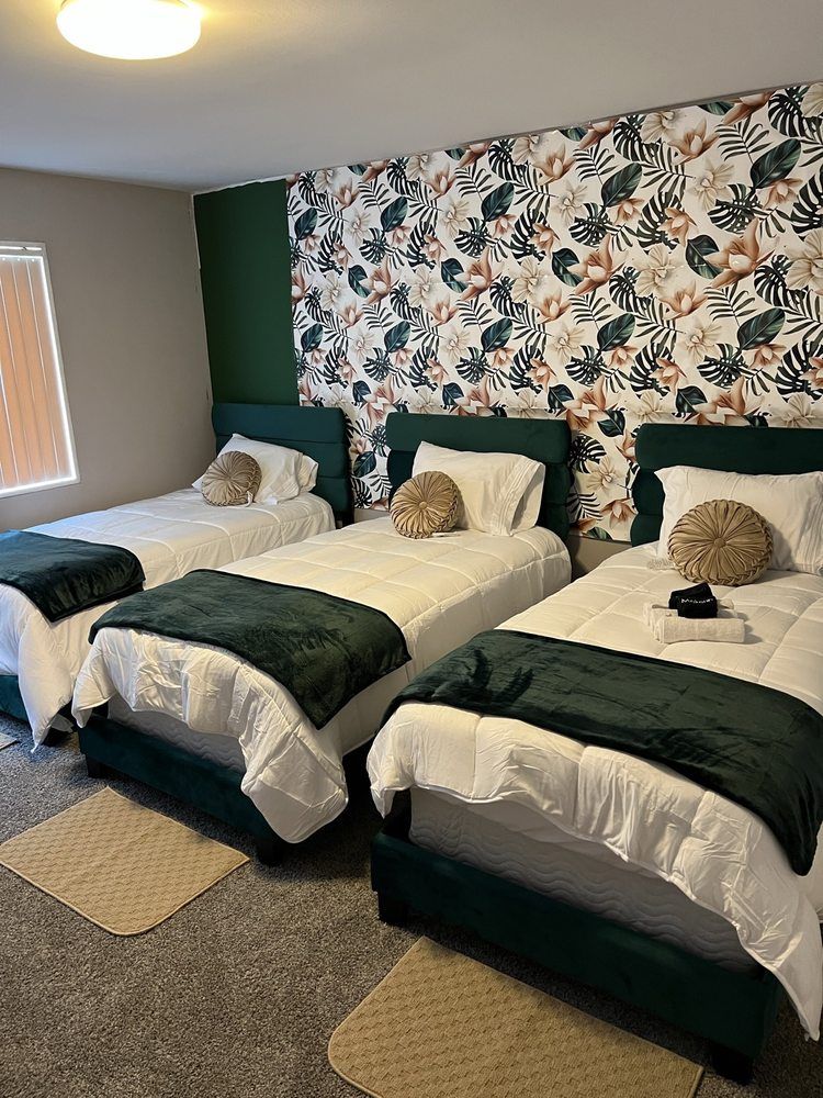Three beds in a room with patterned wallpaper and green accents. Each bed has white bedding and a green throw blanket.