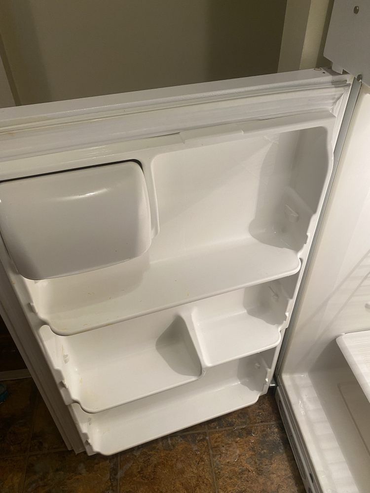 Open white refrigerator door with empty shelves. The interior is white, showing a top compartment and three shelves.