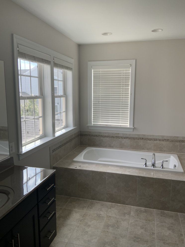 Bathroom with a large bathtub, surrounded by tile, under a window with blinds. Vanity and windows are visible as well.