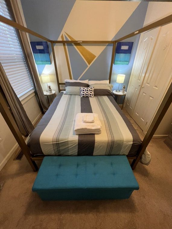 Bedroom with a bed frame, striped bedding, and a blue storage bench. A geometric mural is on the wall.
