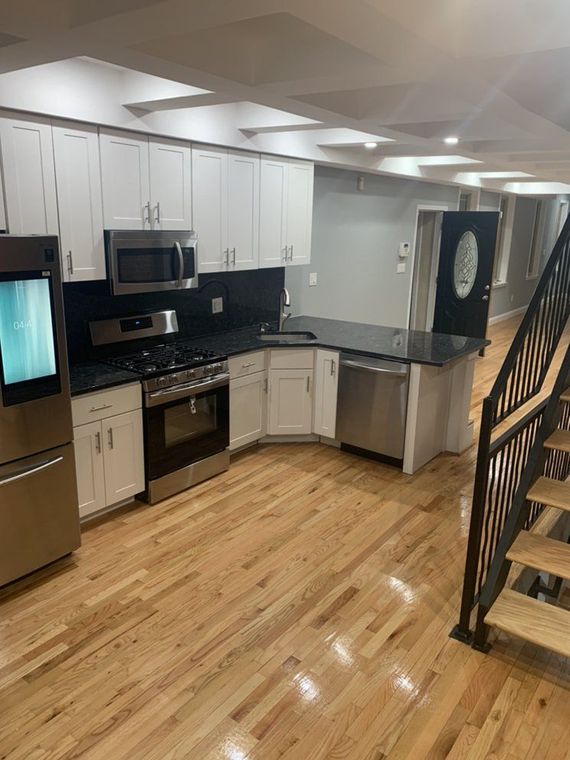 Modern kitchen with white cabinets, stainless steel appliances, dark countertops, and hardwood floors. Stairs lead upward.