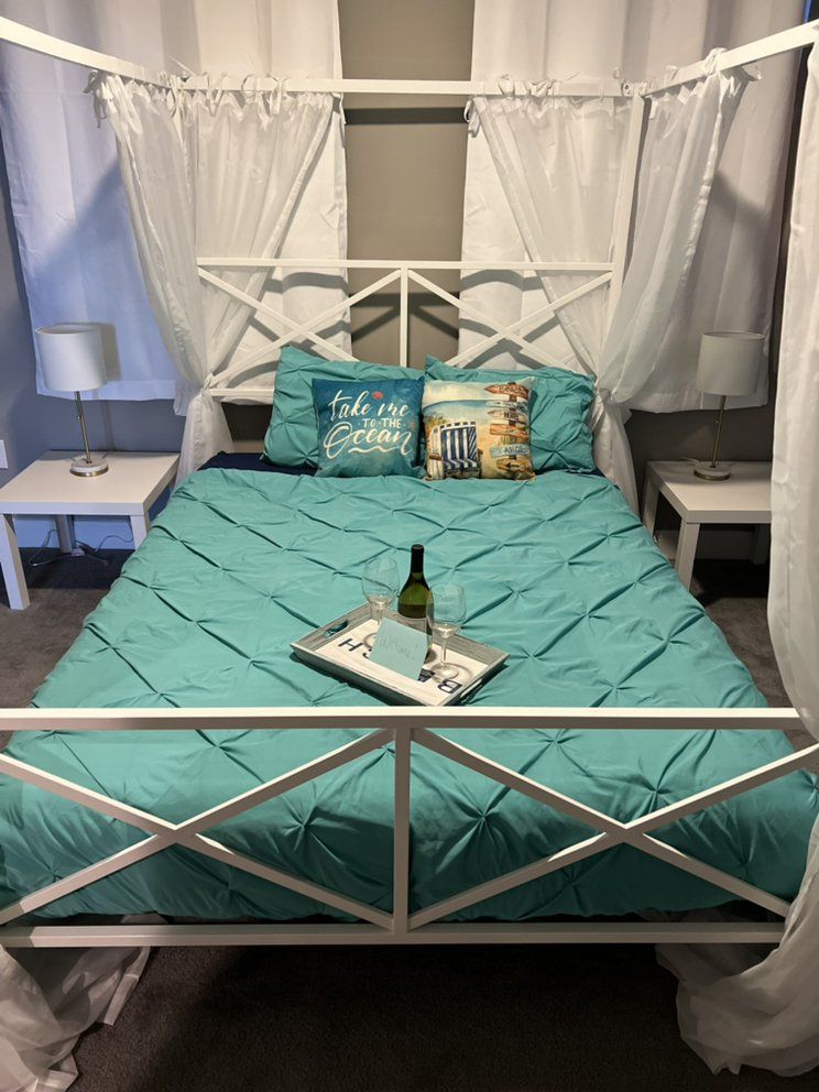 A white canopy bed with turquoise bedding and pillows. A tray with a bottle of wine sits on the bed.