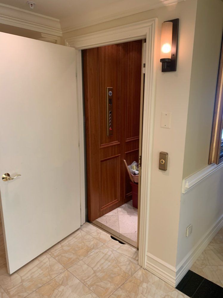 Open doorway revealing a wood-paneled elevator.  White door on the left, light fixture and elevator controls on the right.