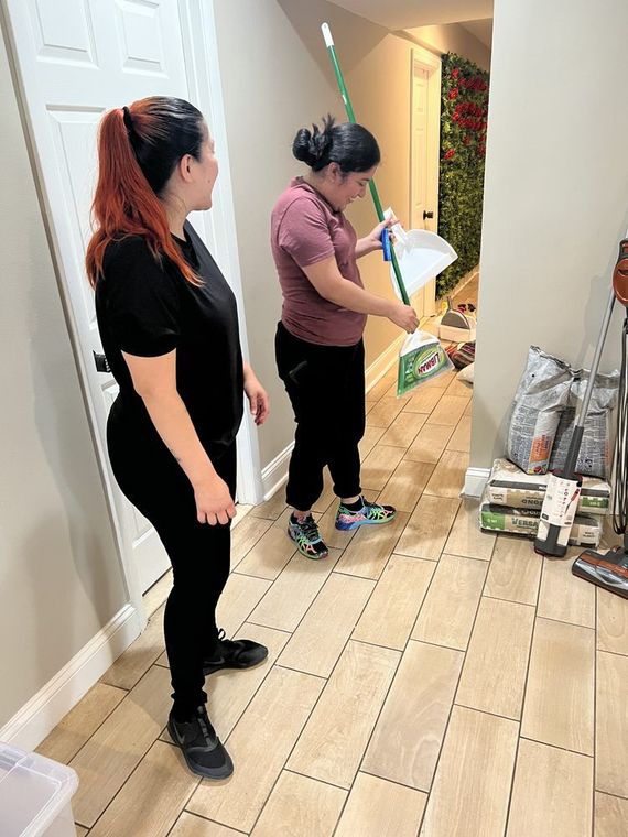 Two women cleaning a hallway. One sweeps debris into a dustpan, while the other watches.