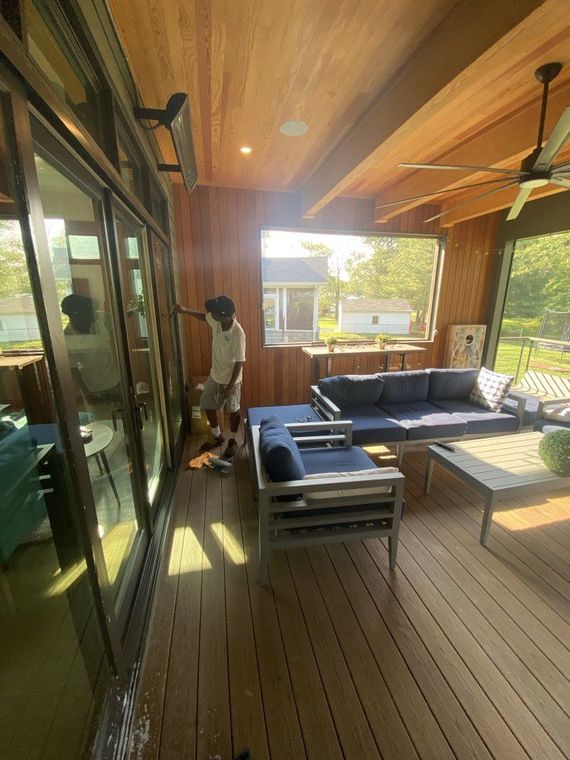 A person standing near glass sliding doors in a modern, wooden patio with blue couches and a table. Sunny day.