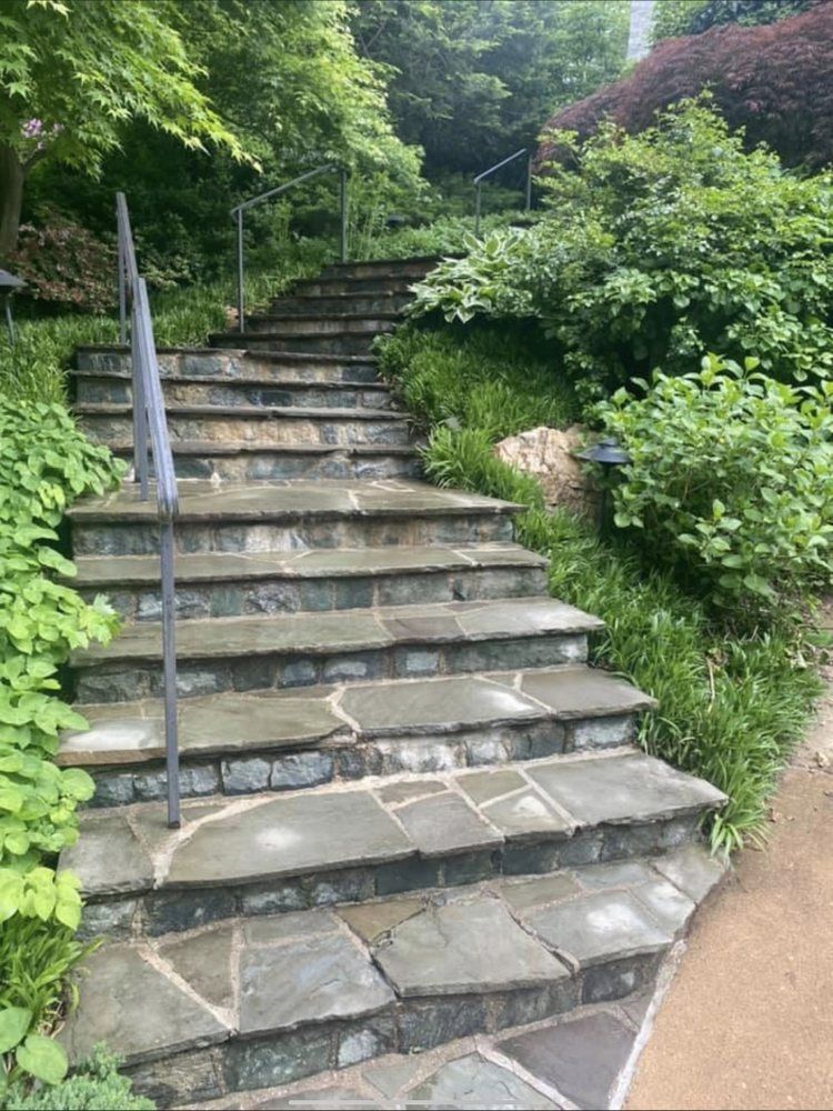 Stone steps with a metal handrail ascend a lush, green hillside garden.