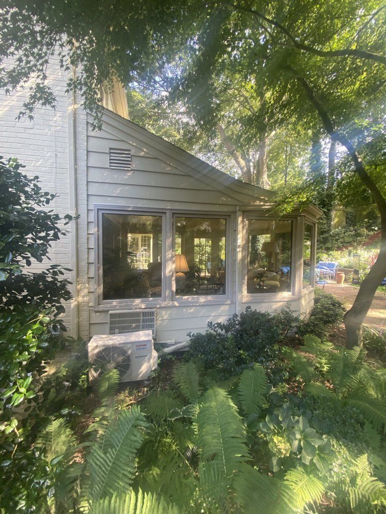 Exterior view of a light-colored house with a row of windows. A/C unit sits below, surrounded by greenery and trees.