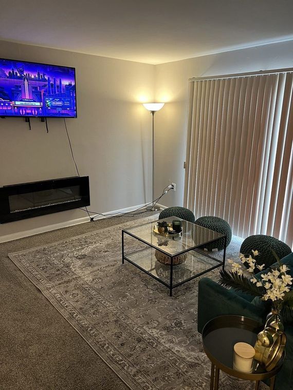 Living room with a mounted TV, electric fireplace, gray rug, and teal furniture. A glass coffee table sits in the center.