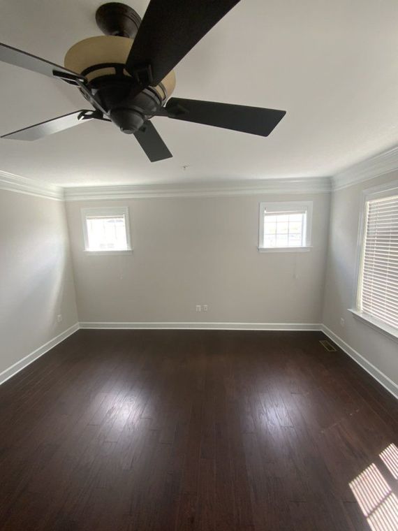 Empty room with dark wood floors, white walls, and two small windows. A ceiling fan hangs from the white ceiling.