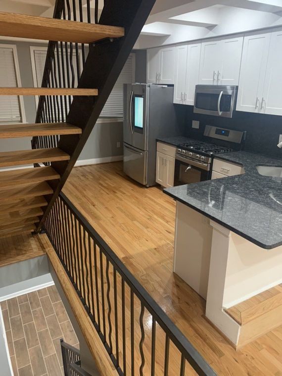 A kitchen with hardwood floors, white cabinets, and a granite countertop next to a set of stairs with a black metal railing.
