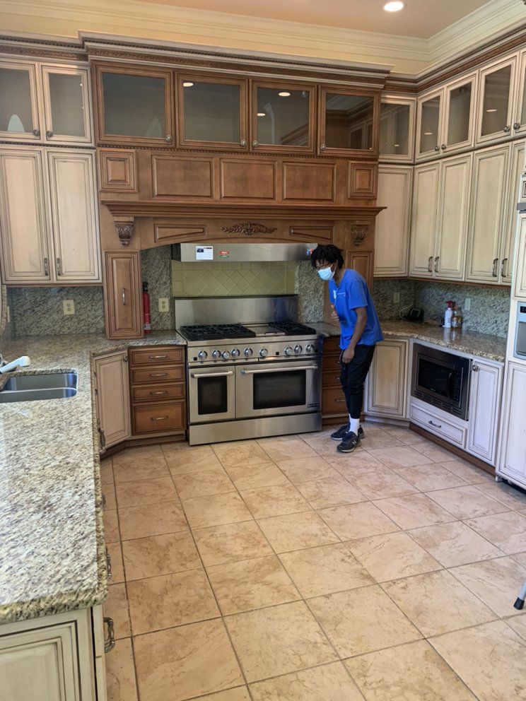 Person in a blue shirt and face mask standing in a large kitchen with stainless steel appliances and wooden cabinets.