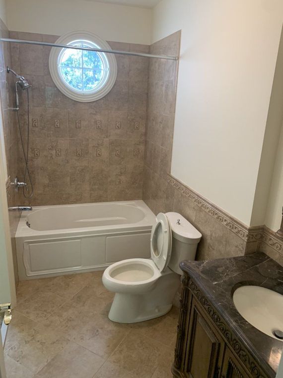 Bathroom with a bathtub, toilet, and vanity. Beige tiles cover the floor and walls. A round window is above the tub.