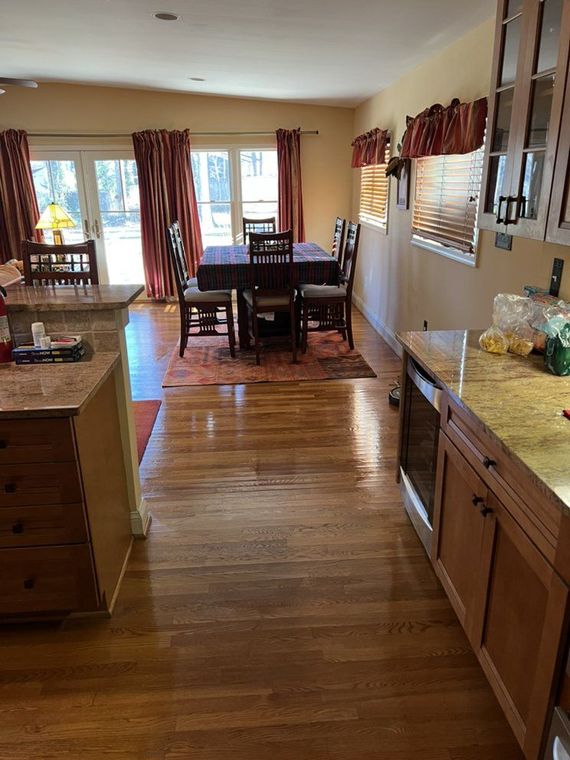 Kitchen with wooden floors leading to a dining area with a table and chairs. Red curtains frame the windows.