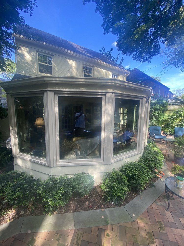 A two-story beige house with large bay windows, surrounded by greenery and a brick patio.