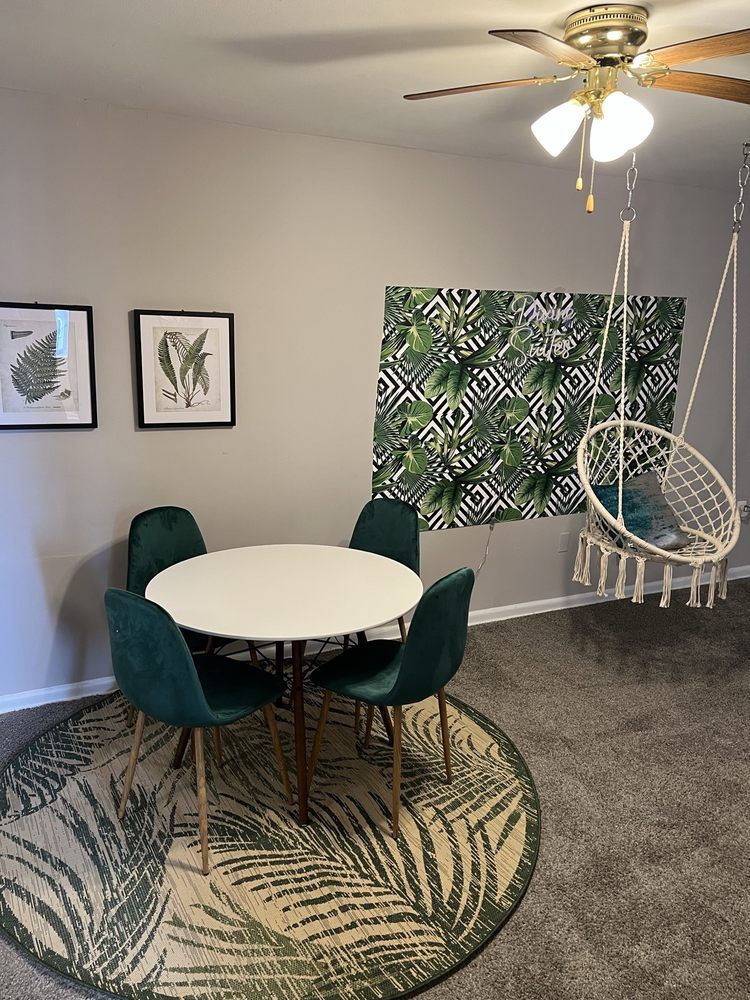 A dining area with a white table, four teal chairs, and a tropical leaf rug. A swing chair hangs nearby.