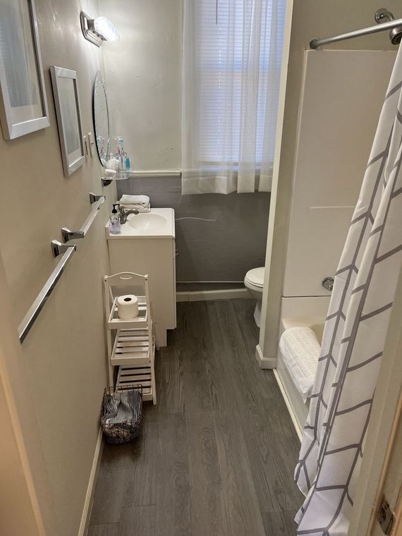 A narrow bathroom with gray wood-look flooring, a white vanity, and a shower with a patterned curtain.