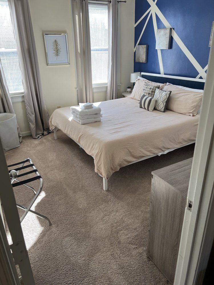 Bedroom with bed, dresser, and geometric blue wall accent. Beige carpet and curtains, sunlight streams in the windows.