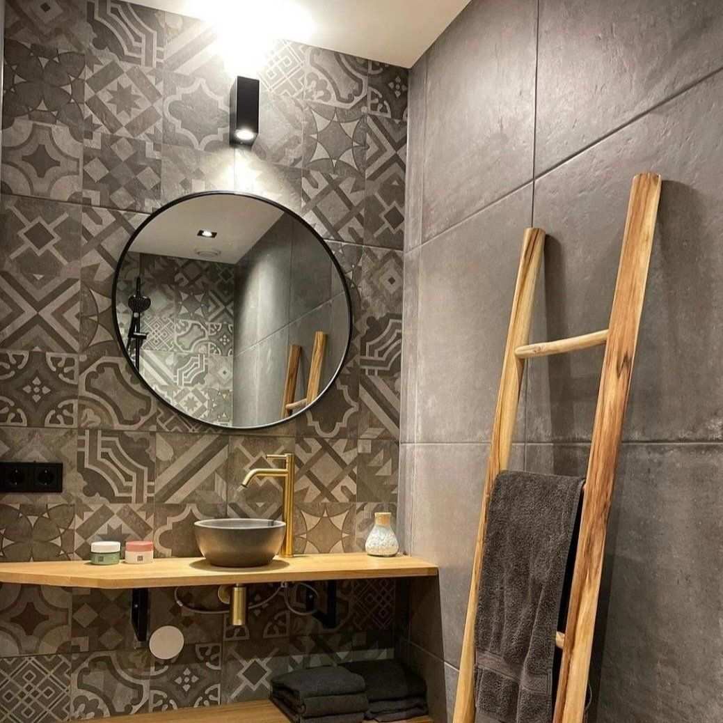 Badkamer met geblokte tegels, ronde spiegel, houten wastafel, ladder en handdoeken; grijs- en bruintinten.