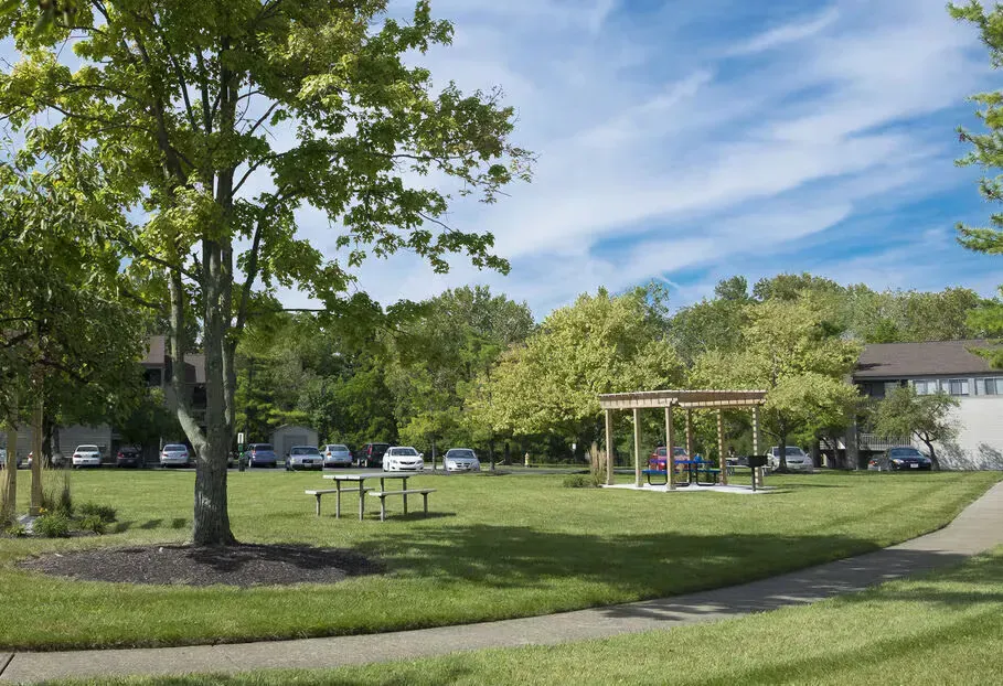 Open grassy courtyard with trees, a gazebo, a picnic table, and a curved sidewalk in an apartment community.