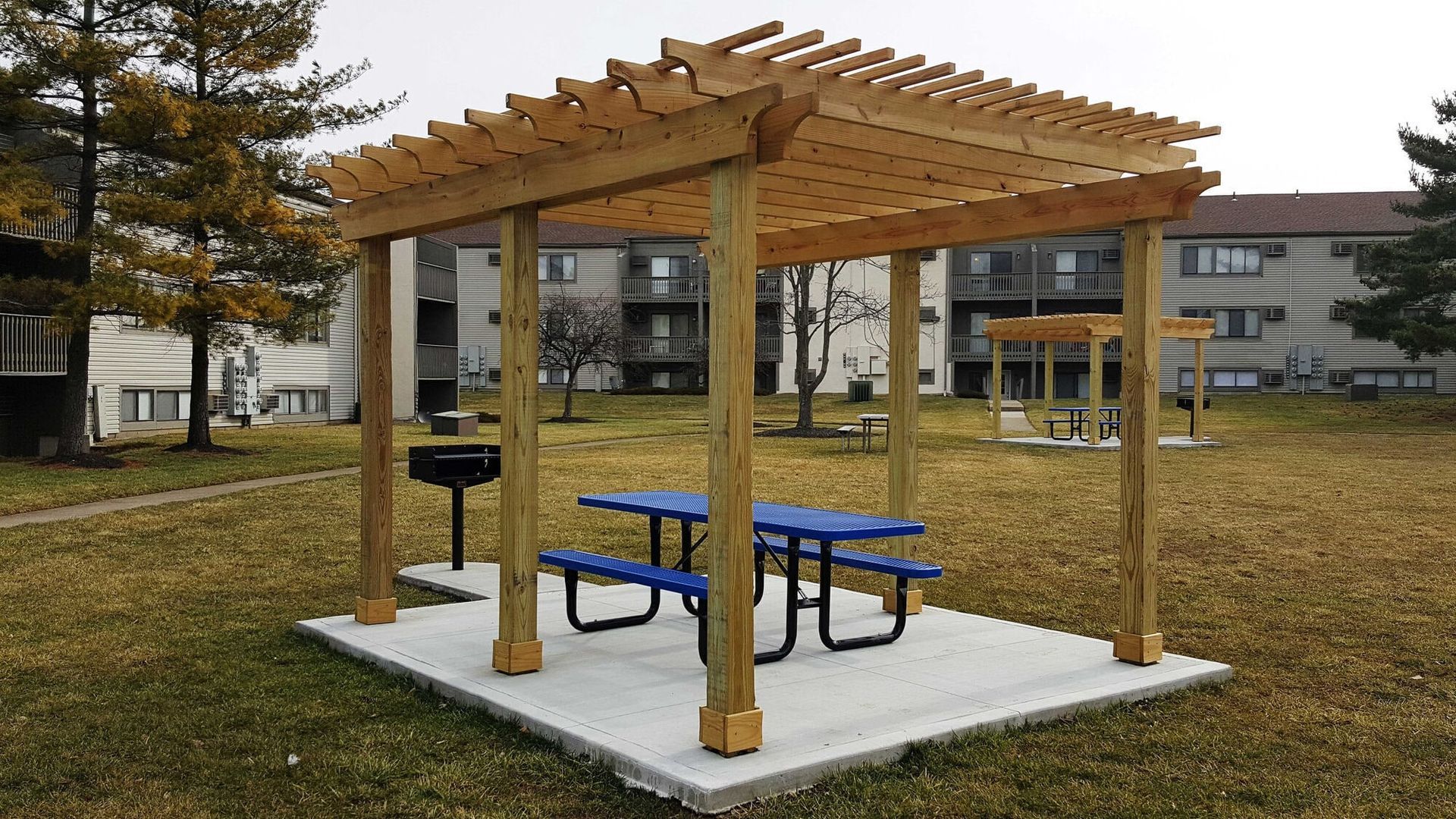 Wood pergola over a concrete pad with a blue picnic table and grill in a courtyard.