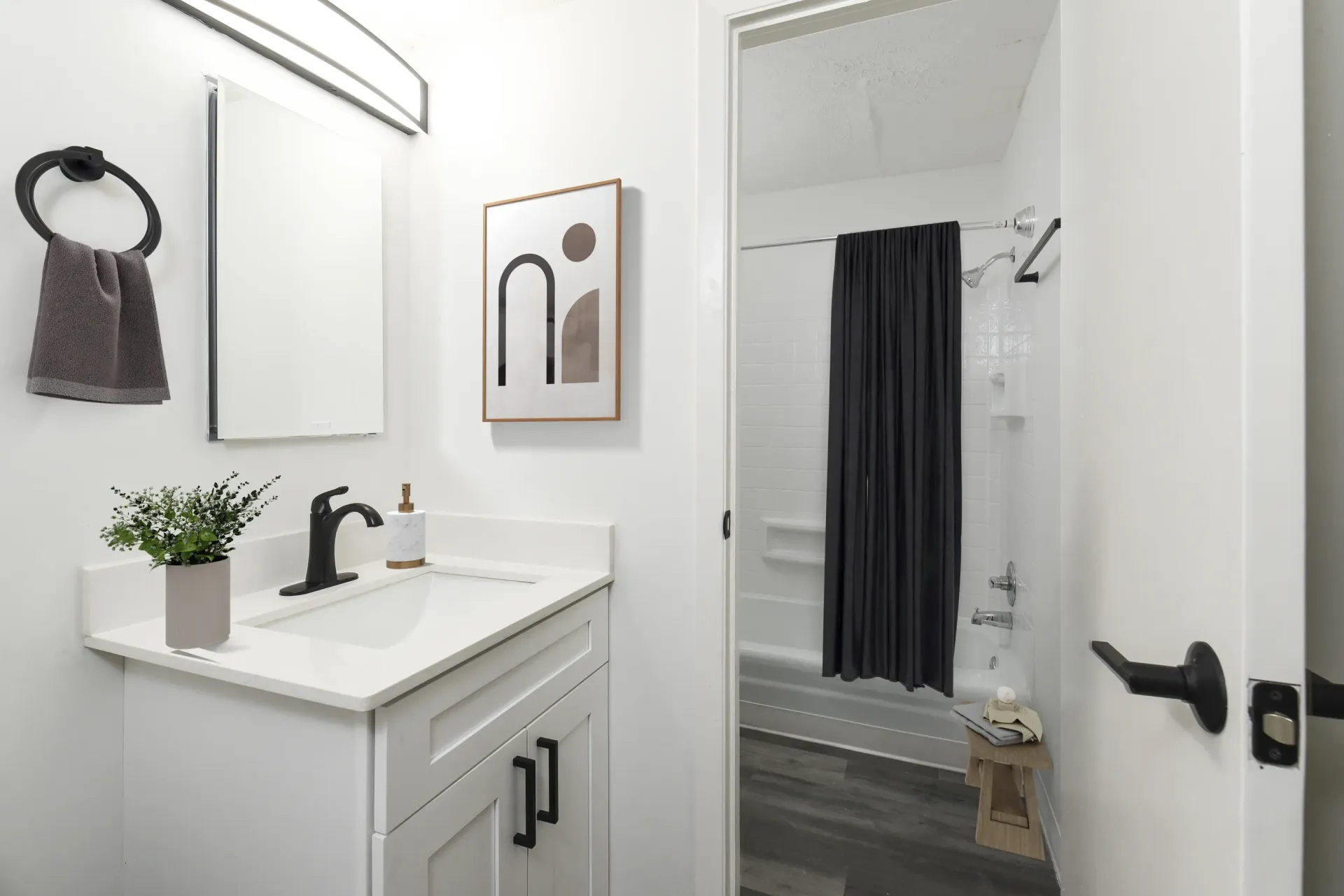 White bathroom with vanity, black faucet, towel ring, framed art, and a shower with a dark curtain.