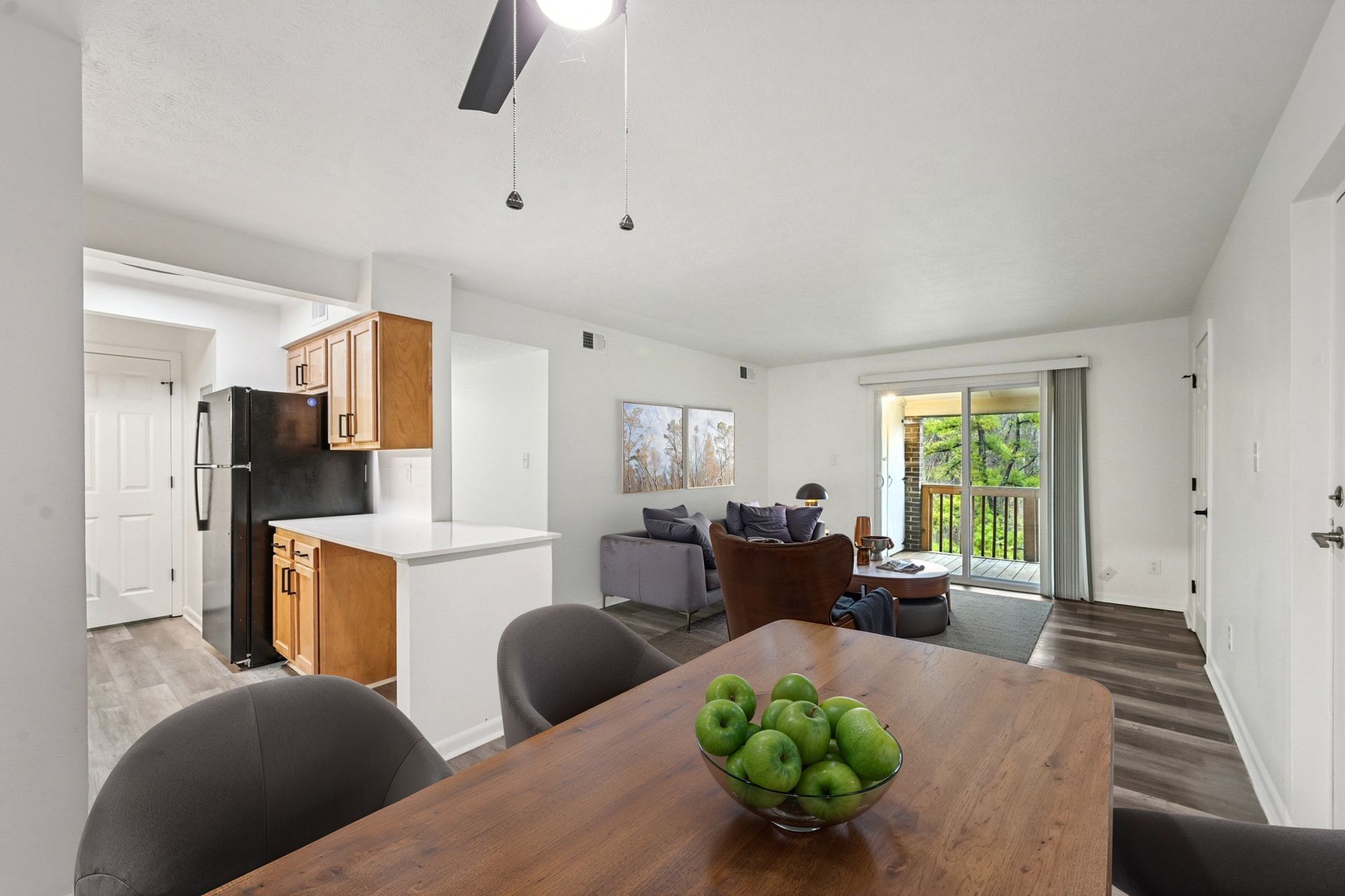 Open-concept apartment interior: dining table in foreground, kitchen to the left, living area and balcony doors.