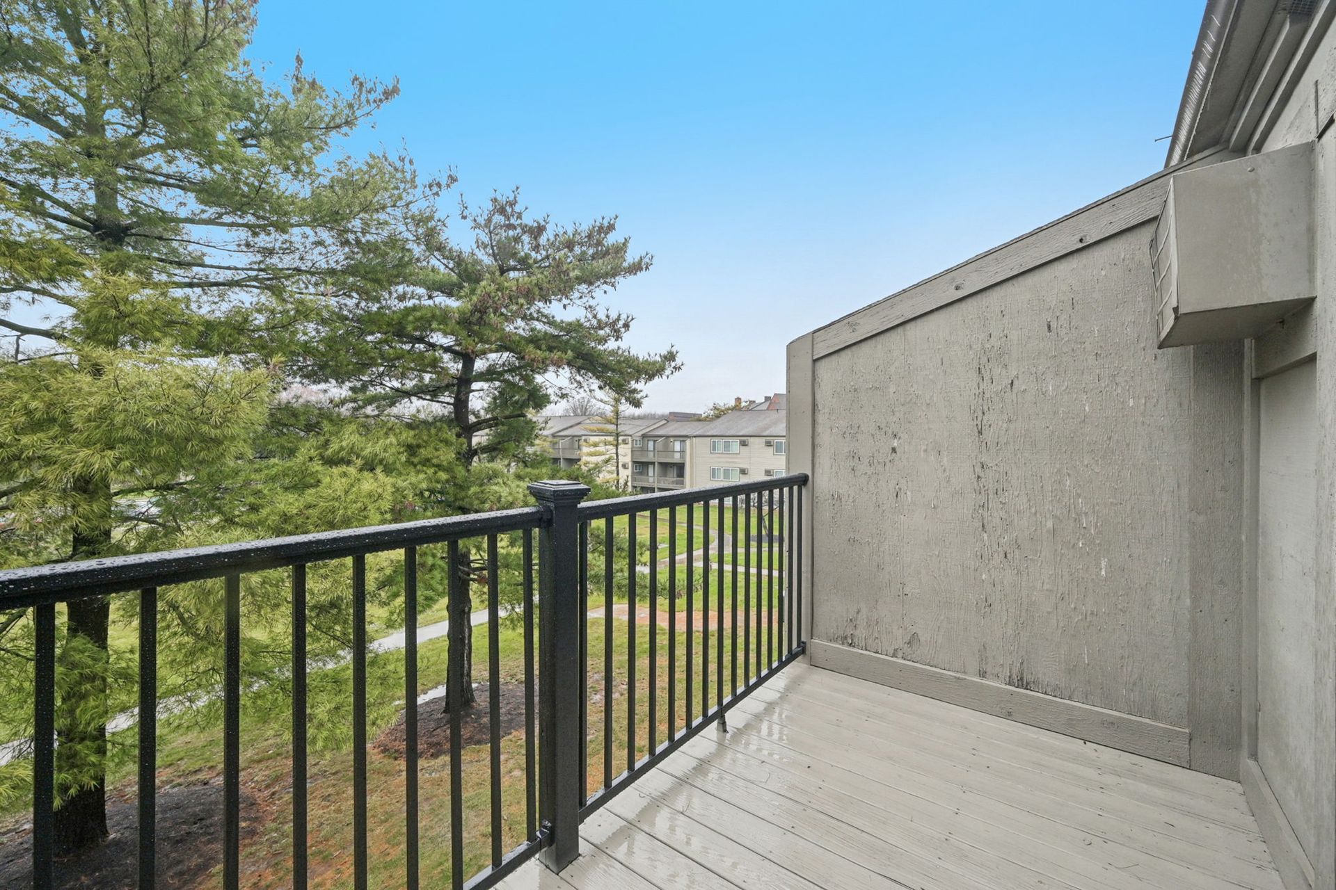 Balcony with black metal railing and light gray wooden deck overlooking trees and neighboring buildings.