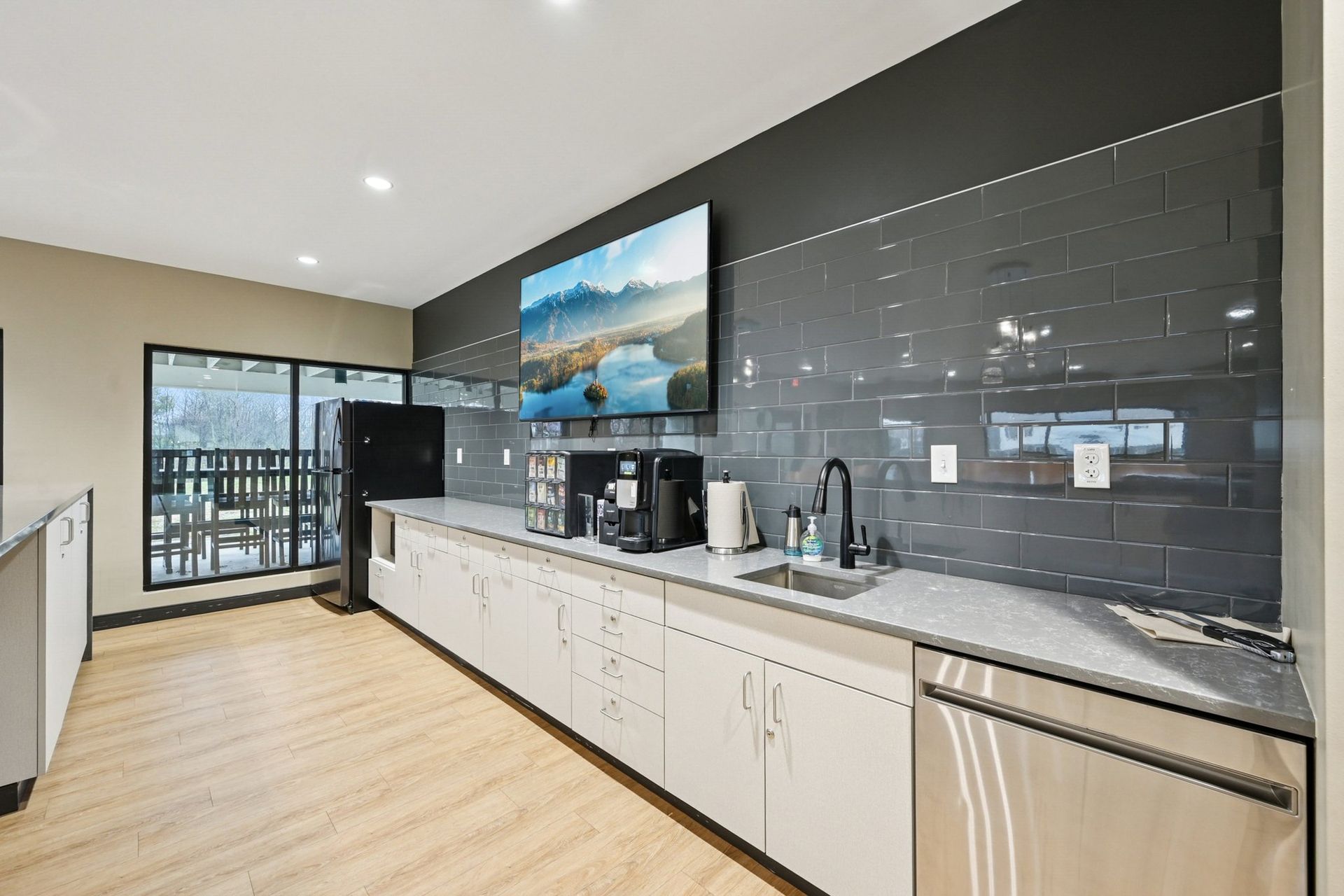 Modern communal kitchenette with white cabinets, gray counters, dark tile backsplash, a mounted TV, and a sliding door.