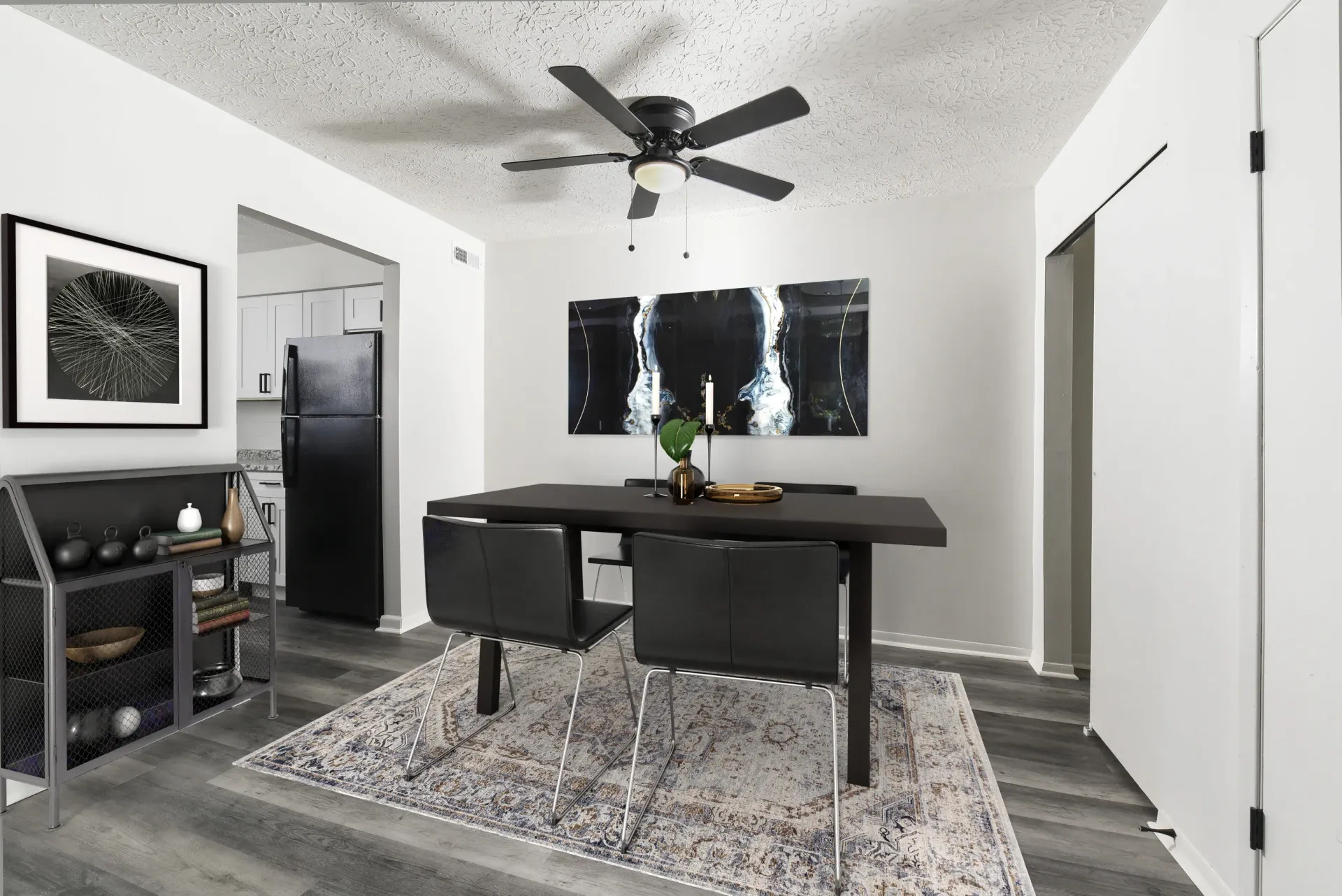 Dining area in a modern apartment: dark rectangular table, four black chairs, ceiling fan, rug, and wall art.