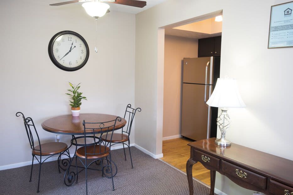 Dining area with round wooden table and metal chairs, clock on the wall, open kitchen with fridge.