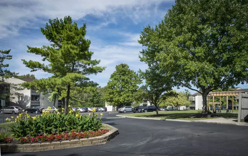 Exterior view of a landscaped apartment community entrance with trees, flowerbeds, and parked cars.