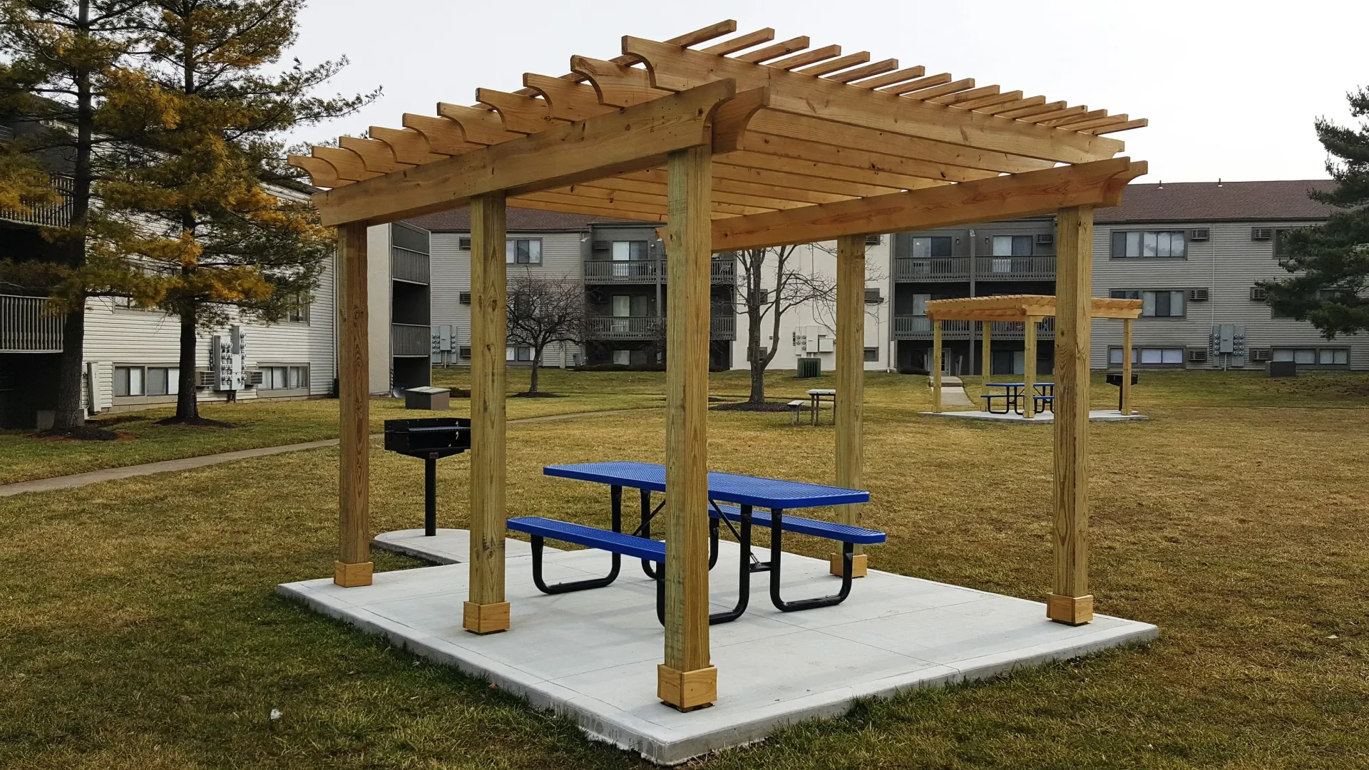 Outdoor communal picnic area with a wooden pergola, blue picnic table, and grill in an apartment complex courtyard.