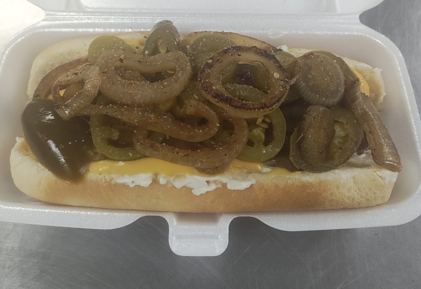 Hot dog in a white container, topped with sauteed onions and jalapeños.