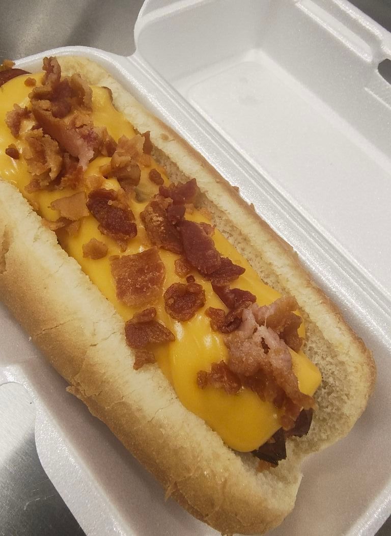 Hot dog in bun, topped with melted cheese and bacon bits, in a white styrofoam container.