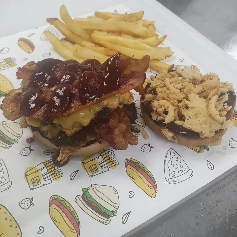 Burger with bacon, cheese, and onion rings, served with fries on a patterned paper.