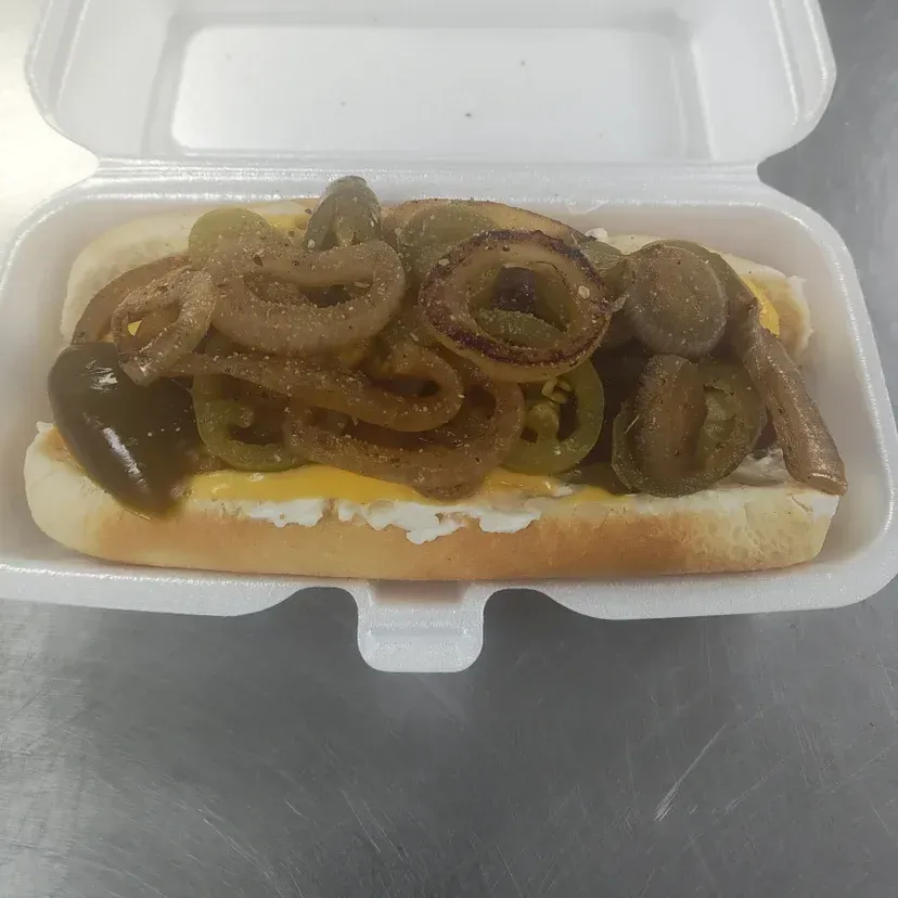 Hot dog in a styrofoam container, topped with fried onions, cheese, and peppers.