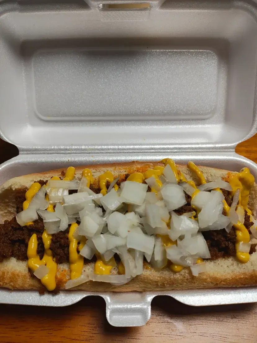 Hot dog in a styrofoam container, topped with chili, mustard, and diced onions.