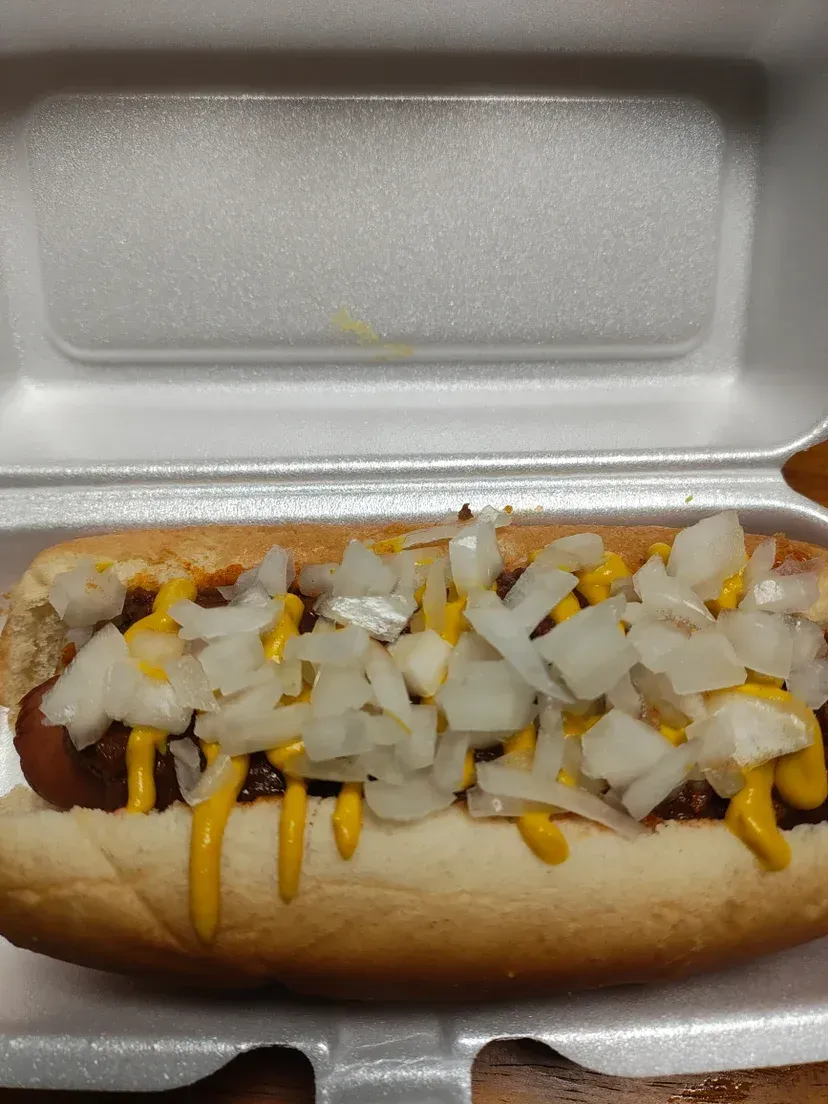 Hot dog in a styrofoam container, topped with chili, mustard, and chopped onions.