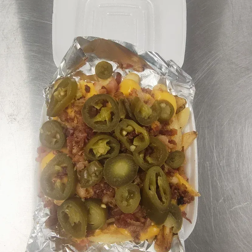 Fries with bacon, cheese, and jalapenos in a white takeout container lined with foil.