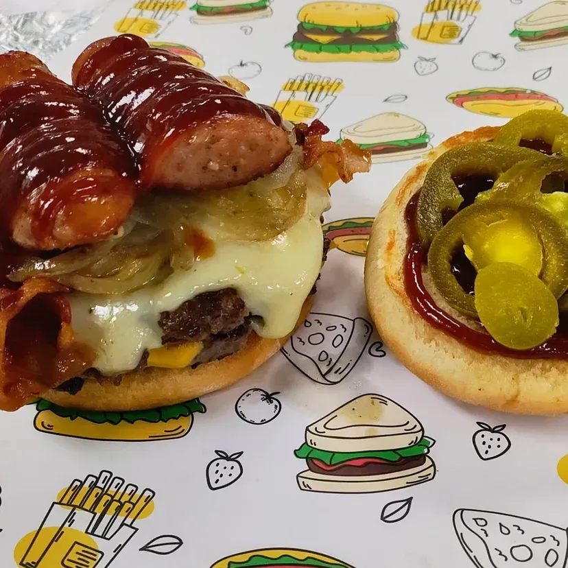 Burger with sausage, bacon, and jalapenos on a decorated food-themed surface.