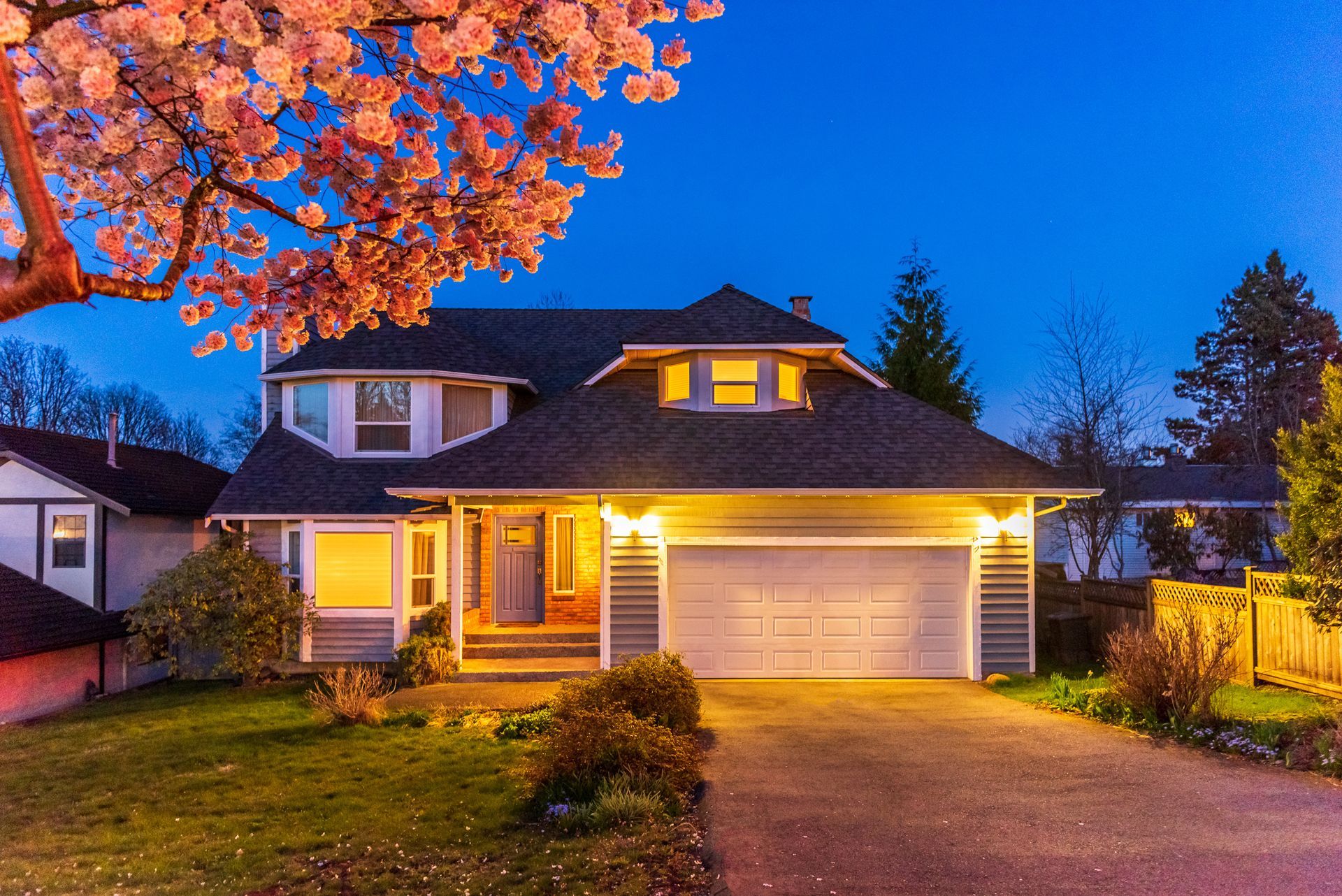 A two-story suburban house with a lit garage at twilight, framed by a blooming cherry tree in the foreground.