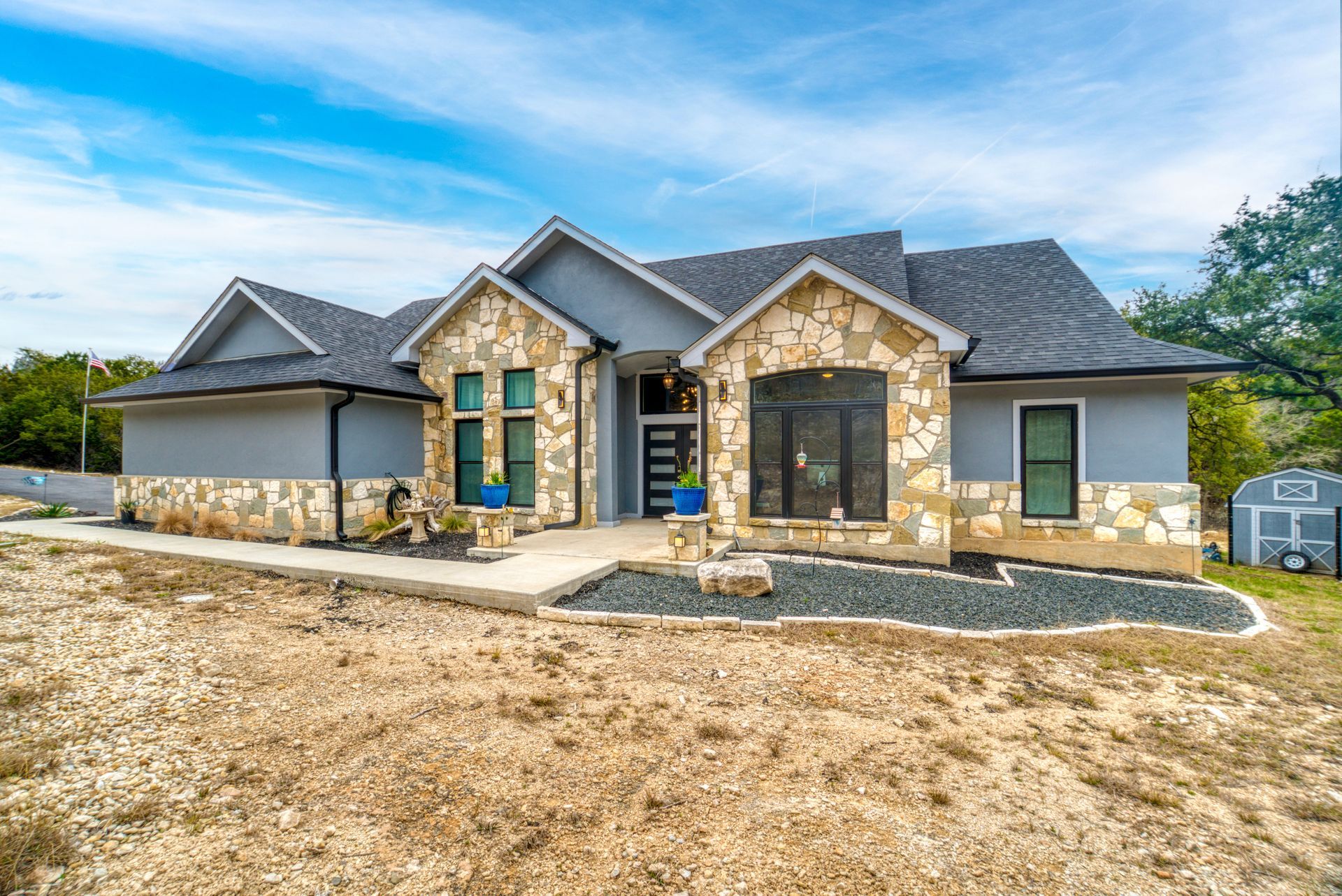 A one-story gray house with stone accents, dark shingles, and a paved walkway, situated on a dry, grassy lot.