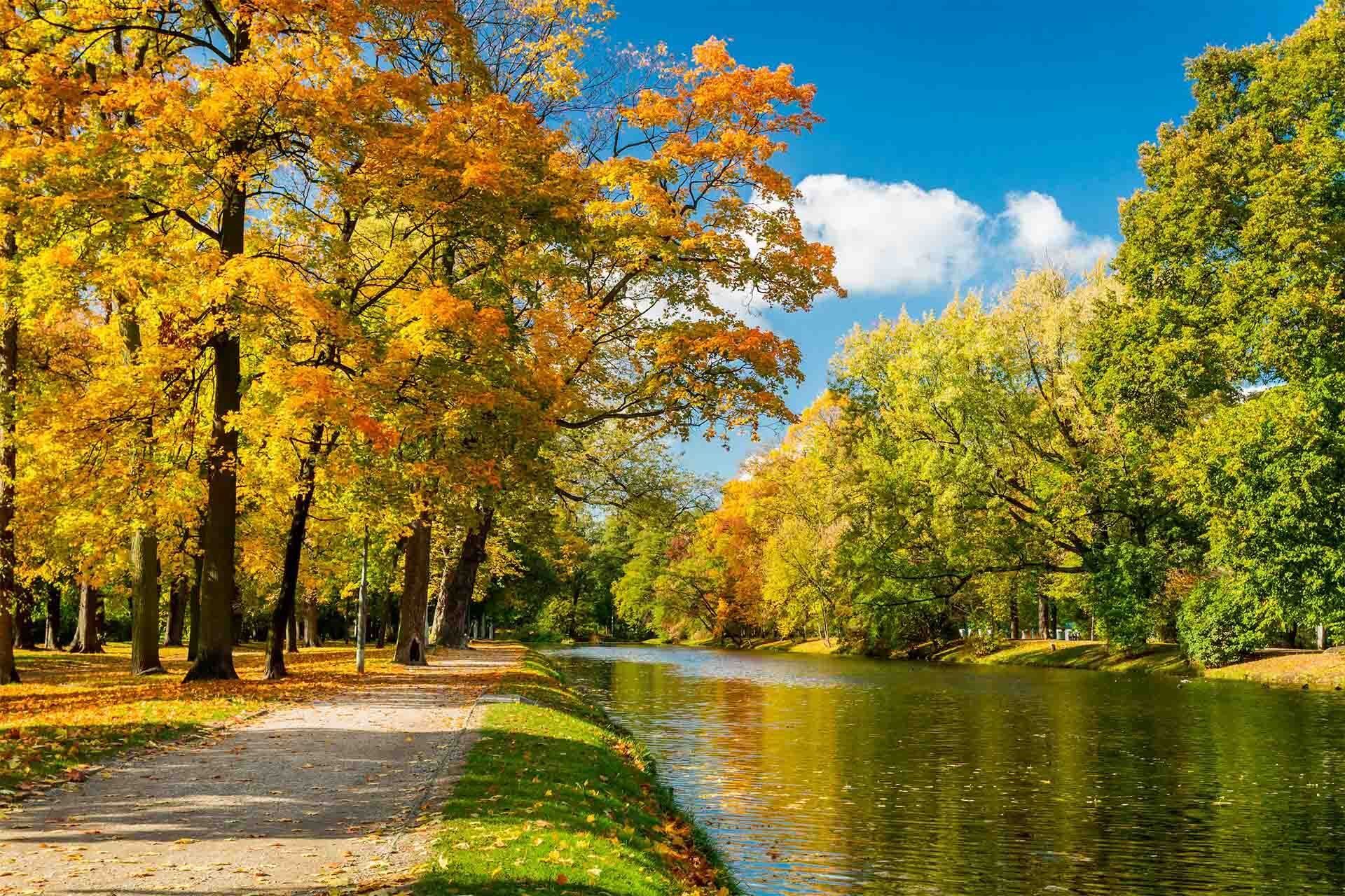 Pathway alongside a canal lined with trees in autumn foliage; blue sky with clouds.